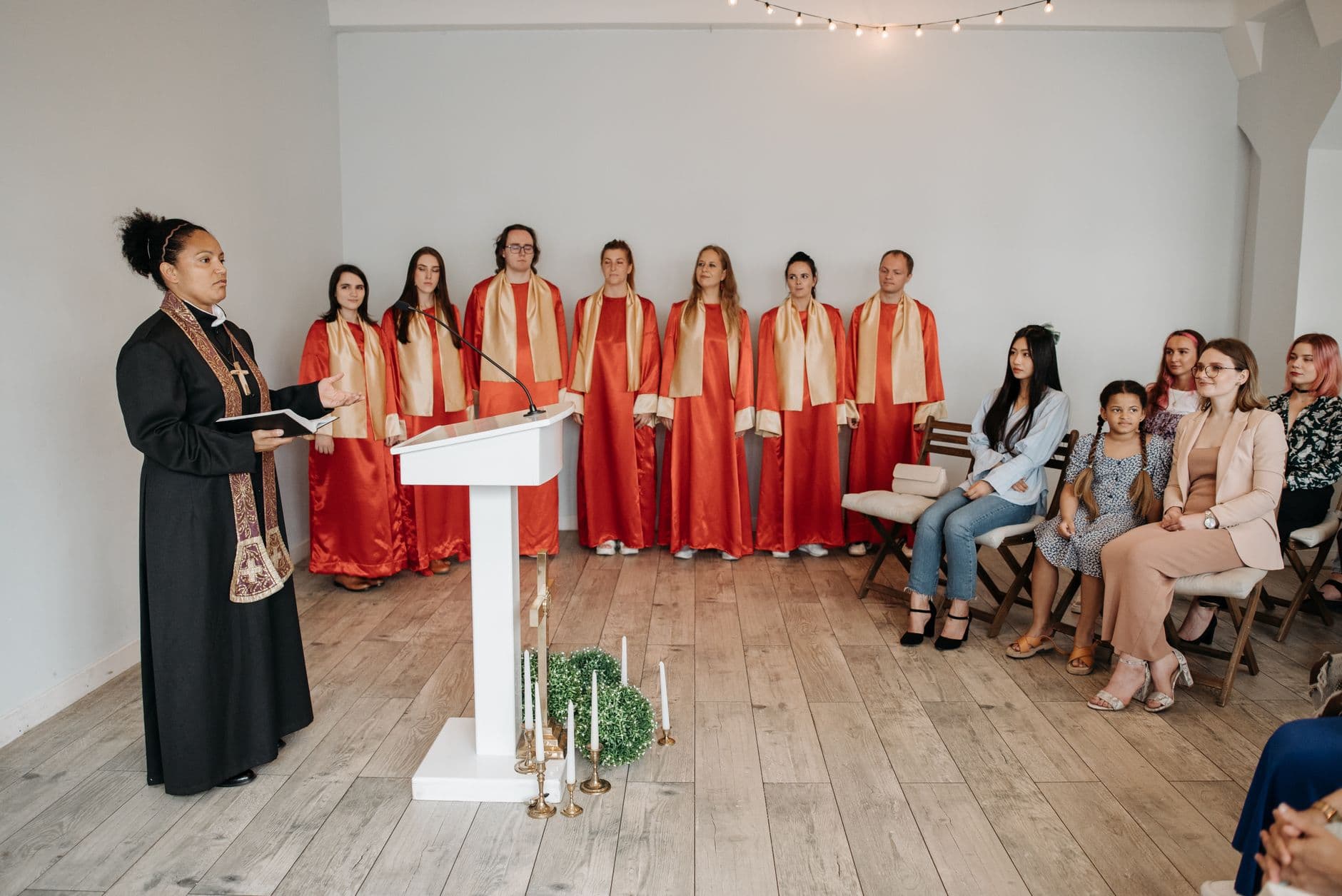 A pastor leads a religious service indoors with a choir in red robes and a seated congregation.